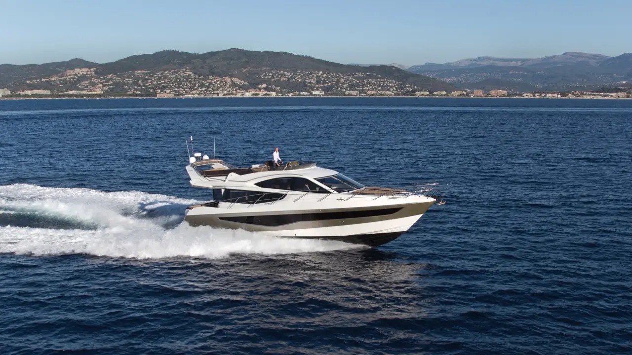 A luxury yacht speeds on deep blue water, with mountains and a coastal city in the background under clear skies.