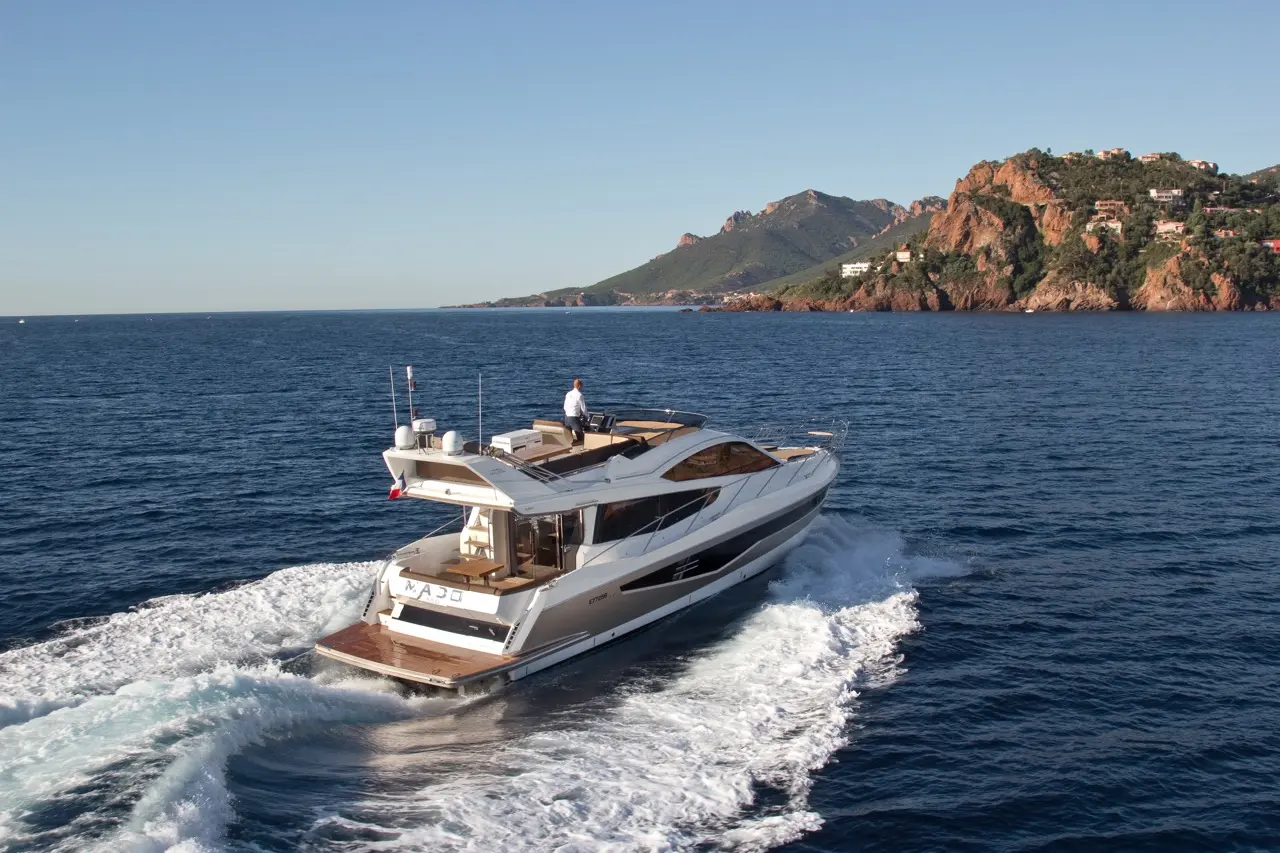 Luxury yacht cruising through the blue sea near rocky hills, with a captain navigating on the deck under clear skies.