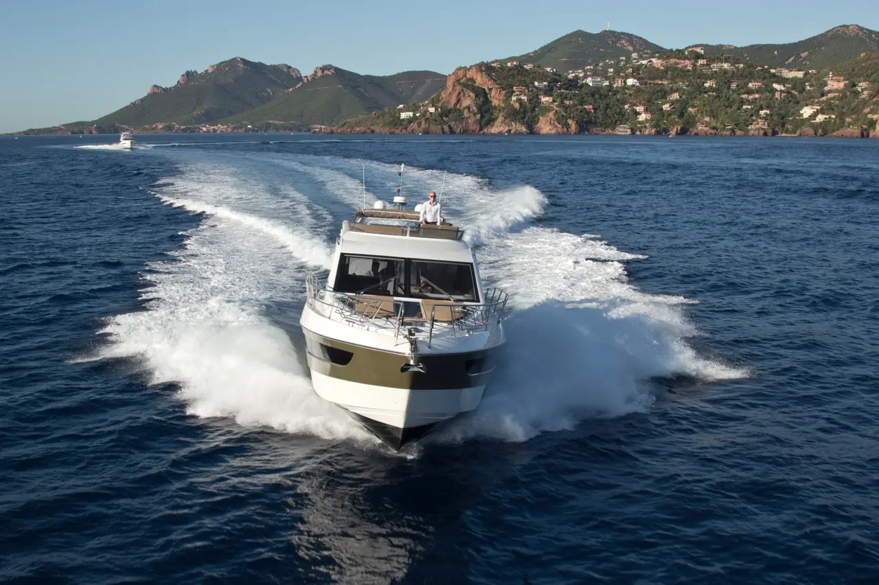 A motorboat speeds through blue waters, creating waves, with mountains and a coastal village visible in the background.