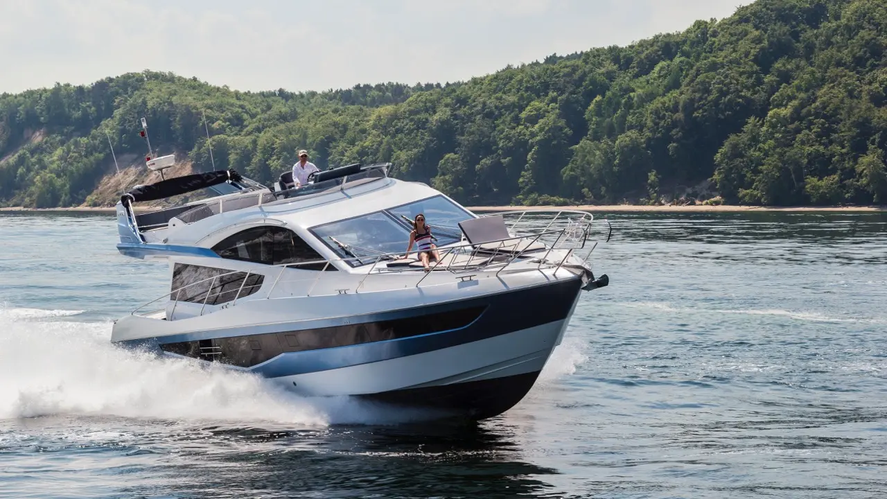 A luxury motor yacht speeds through calm waters, with a woman lounging on the deck and a man at the helm, surrounded by greenery.