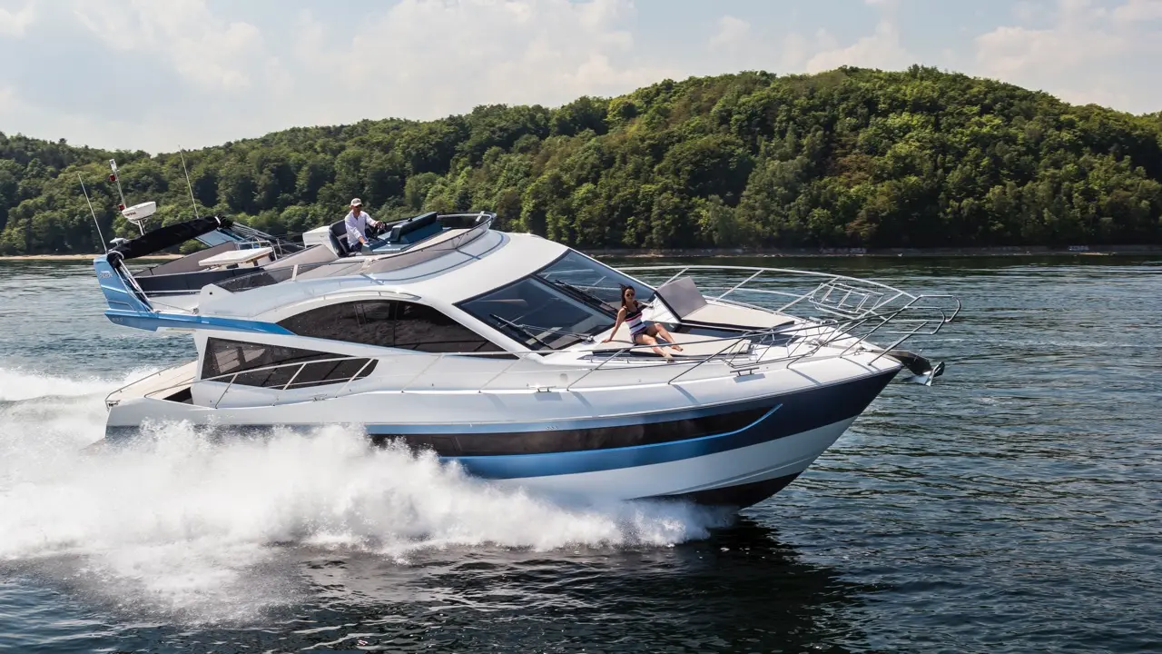 A luxurious motor yacht speeds through water with a woman lounging on the bow and a man steering at the helm.