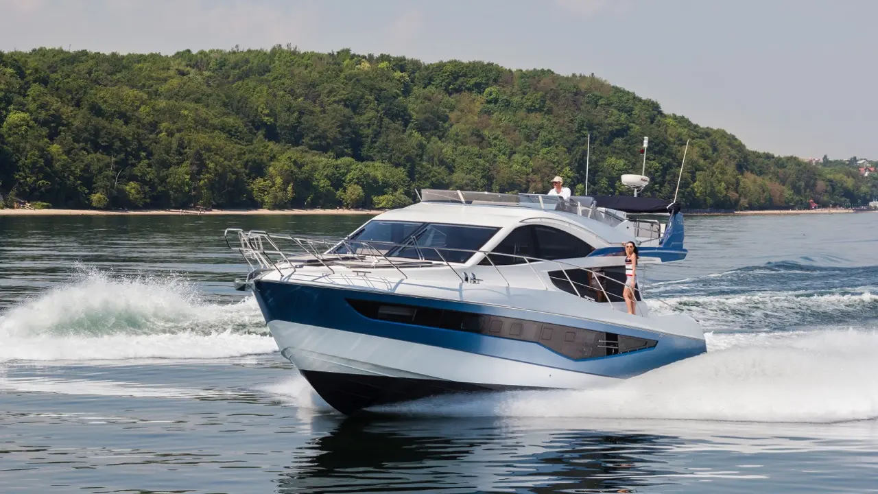 A sleek motor yacht speeds across a calm lake, with a crew member steering and a woman in a swimsuit enjoying the view.
