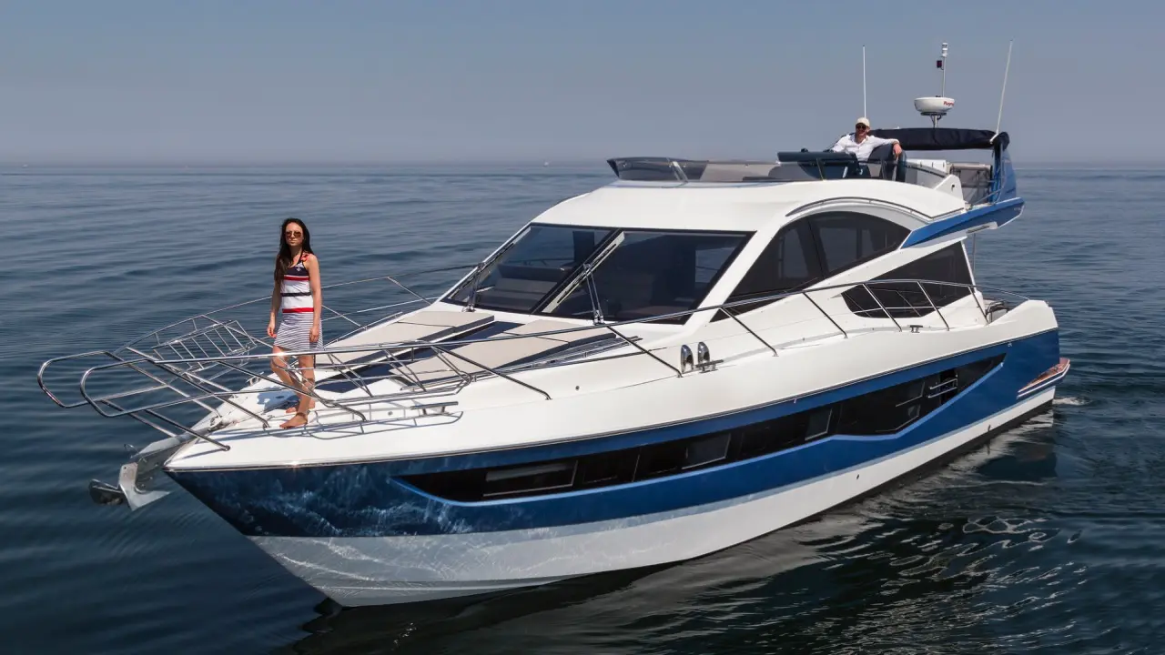 A woman in sunglasses poses on the bow of a luxury yacht, with a male figure relaxing in the background on calm waters.