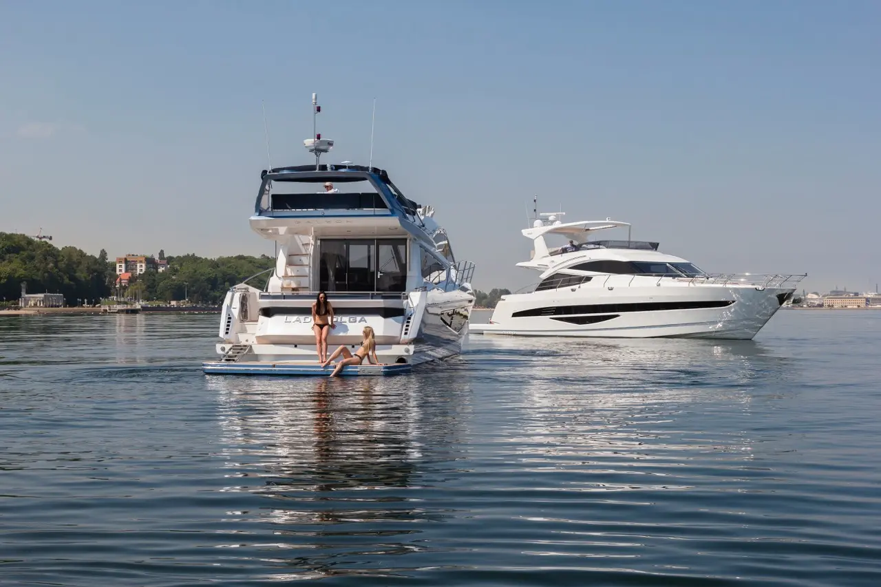 Two women relax on the swim platform of a luxury yacht, with another sleek yacht in the background on calm waters.