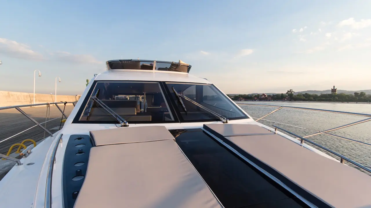 Sunset view of a yacht deck with cozy seating and panoramic windows, waterfront and trees visible in the background.