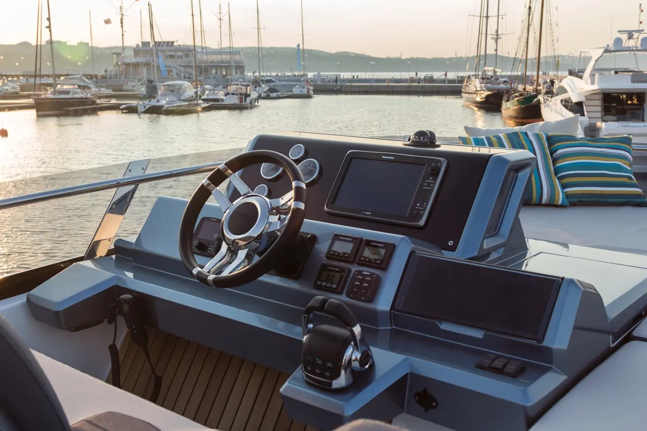 Boat cockpit view with a steering wheel, navigation screen, and controls, set against a harbor at sunset.