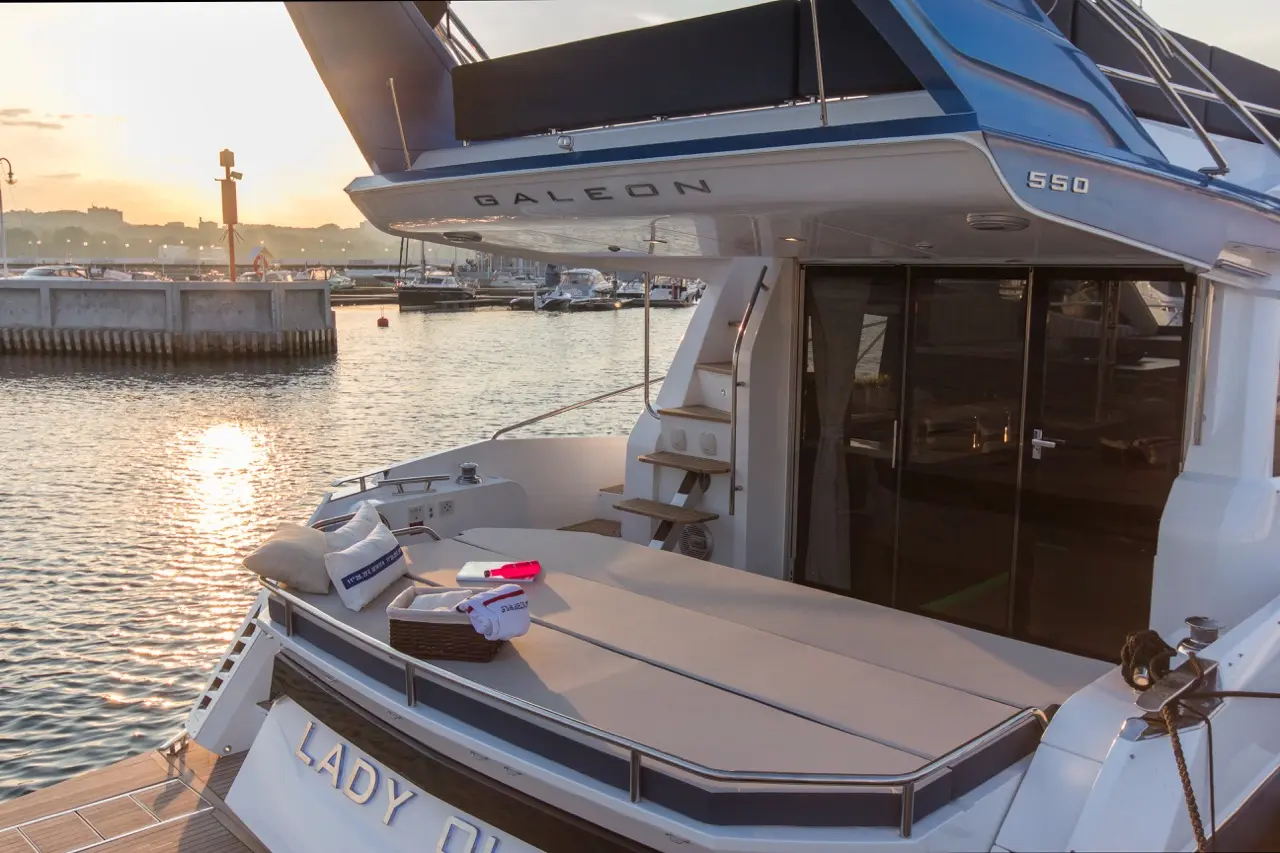 Luxurious Galeon 550 yacht deck with cushions, a wicker basket, and sunset reflection on water in a serene marina setting.