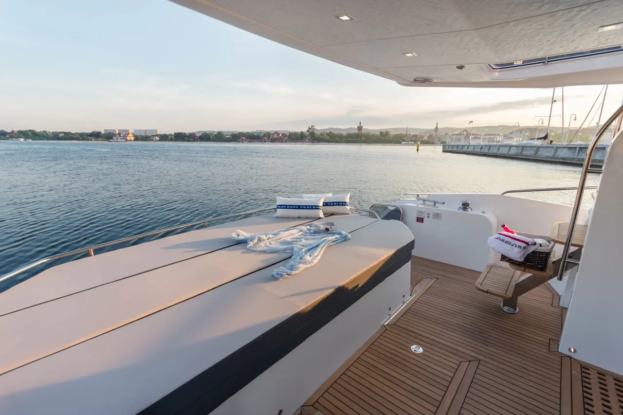 Deck of a yacht with a blue blanket and cushions, overlooking calm water and a distant shoreline during sunset.