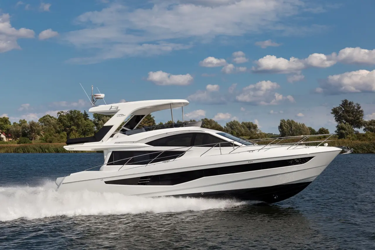 A sleek white yacht cruises through calm waters under a blue sky with scattered clouds and greenery in the background.