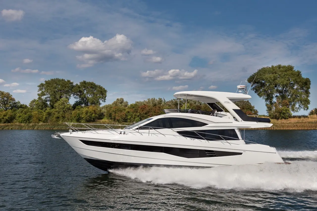 A sleek white yacht cruising on calm water, with greenery in the background and a partly cloudy sky above.