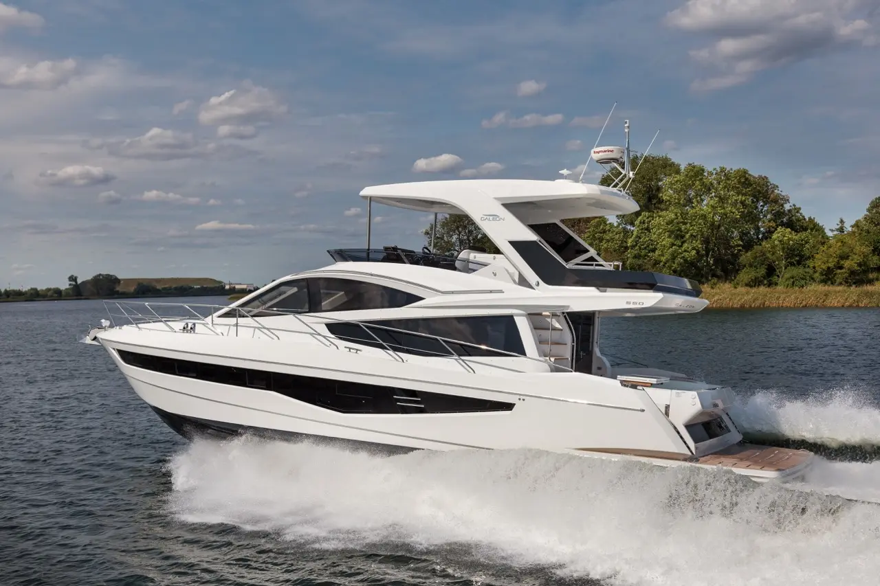 A sleek white yacht cruising over water, creating waves, with a blue sky and green trees in the background.