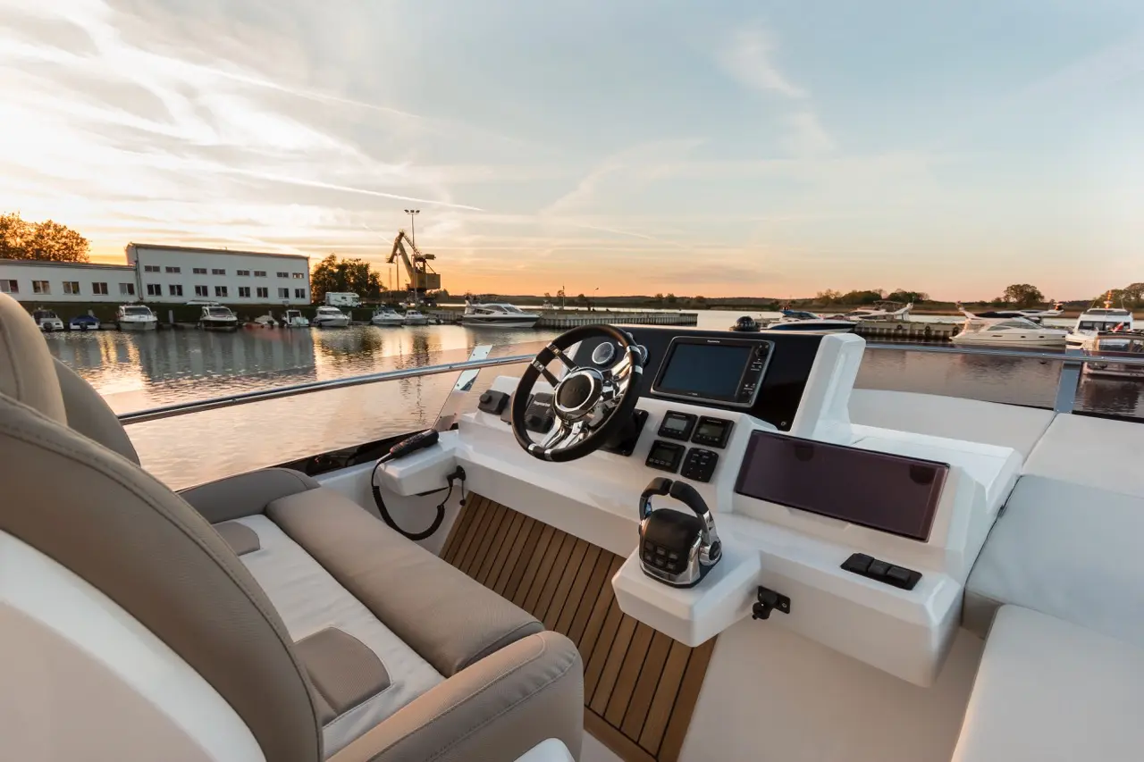 Boat cockpit view at sunset, featuring a steering wheel, control panels, and a serene marina in the background.