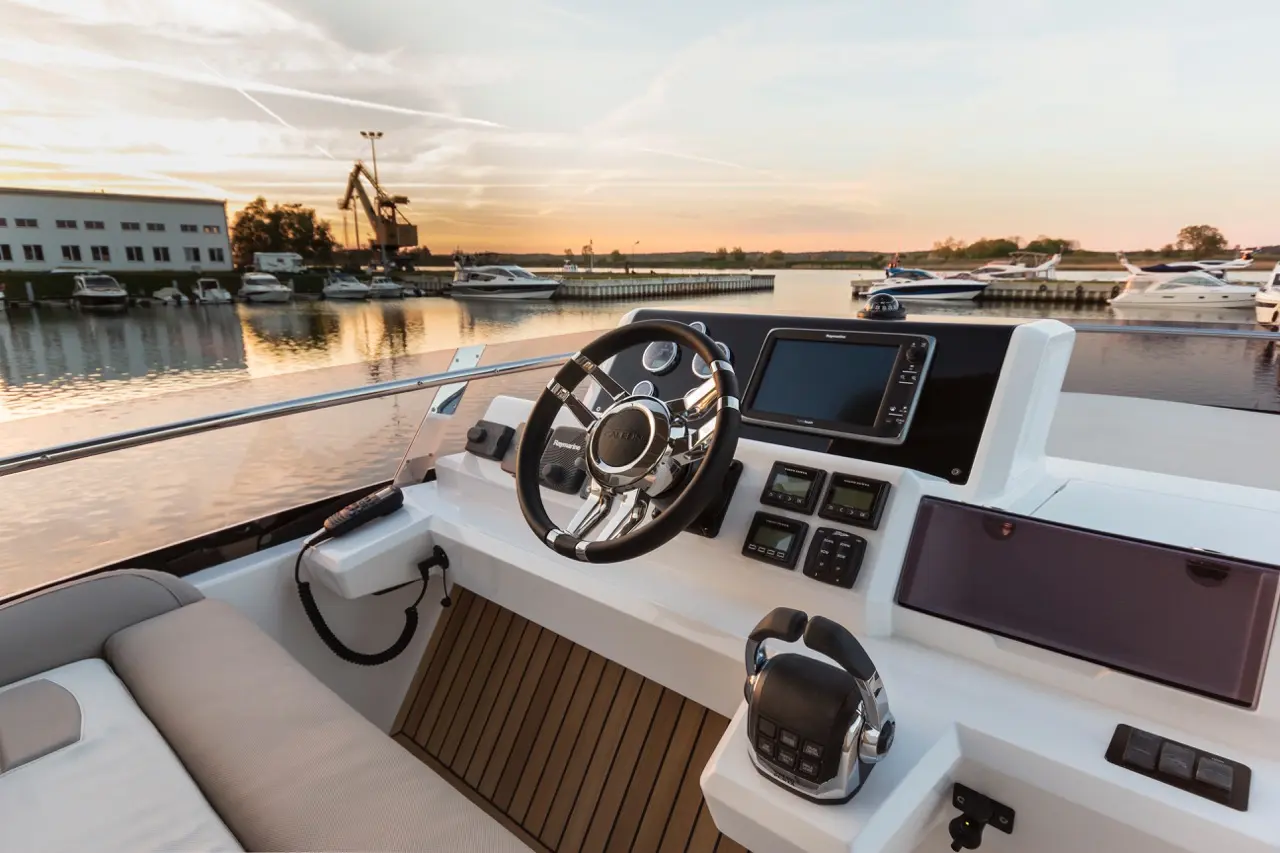 Luxury boat cockpit with a steering wheel, navigation screen, and buttons, overlooking a sunset at the marina.