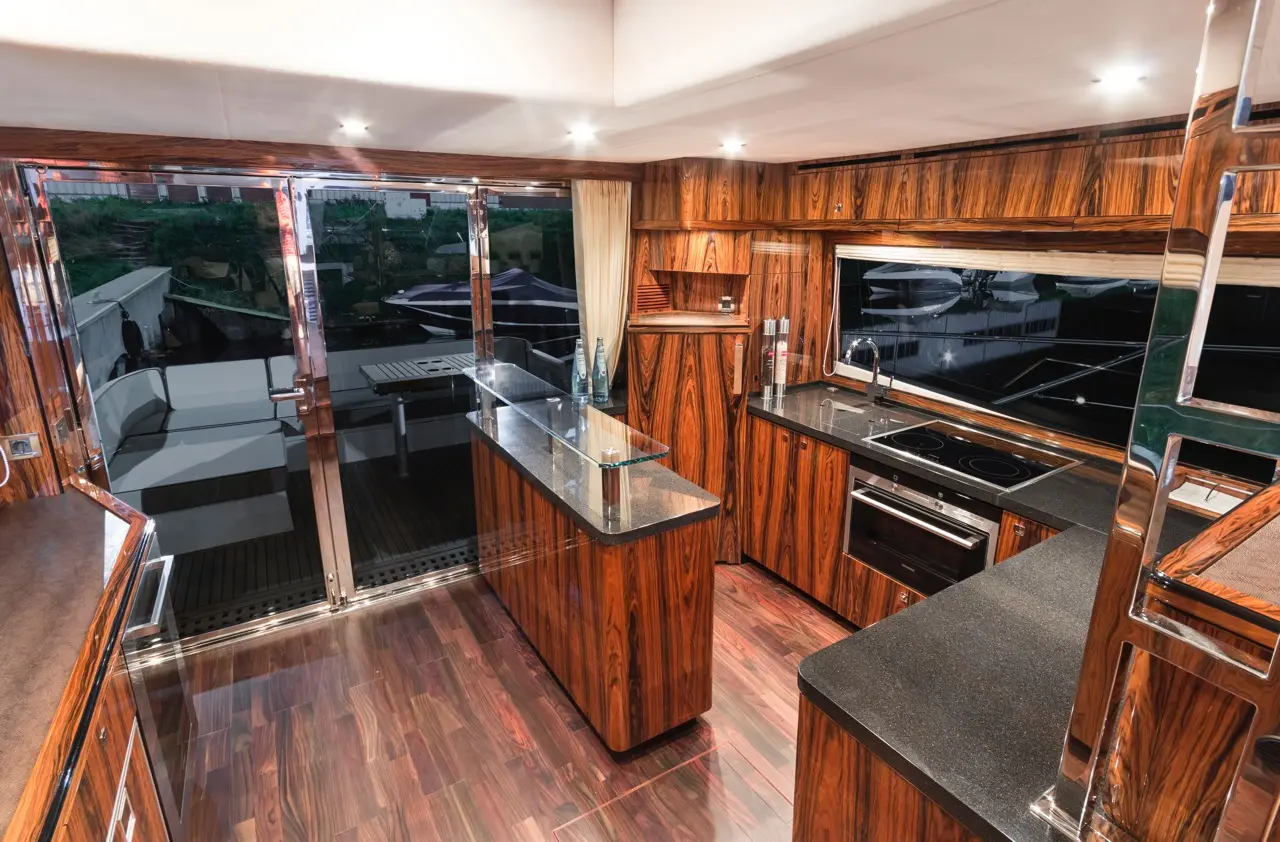 Modern boat kitchen with wooden cabinetry, sleek countertops, and open doors revealing outdoor seating area.