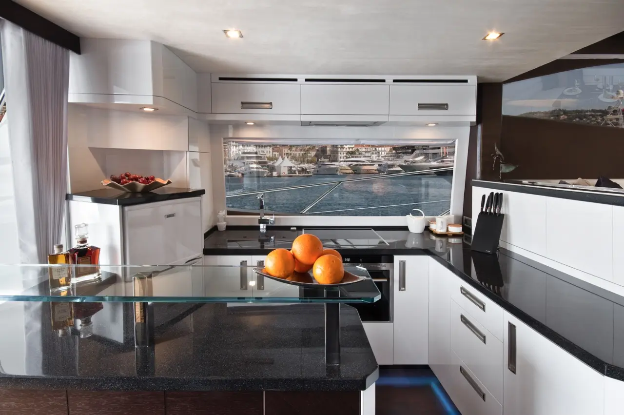 Modern yacht kitchen with sleek white cabinets, black countertops, and a bowl of oranges, offering a view of the marina.