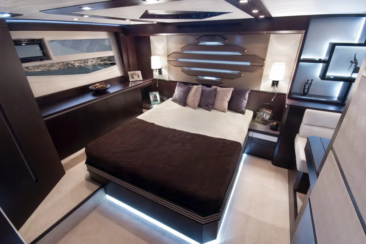 Modern yacht interior featuring a stylish bed with purple pillows, sleek cabinetry, and a view through a large window.