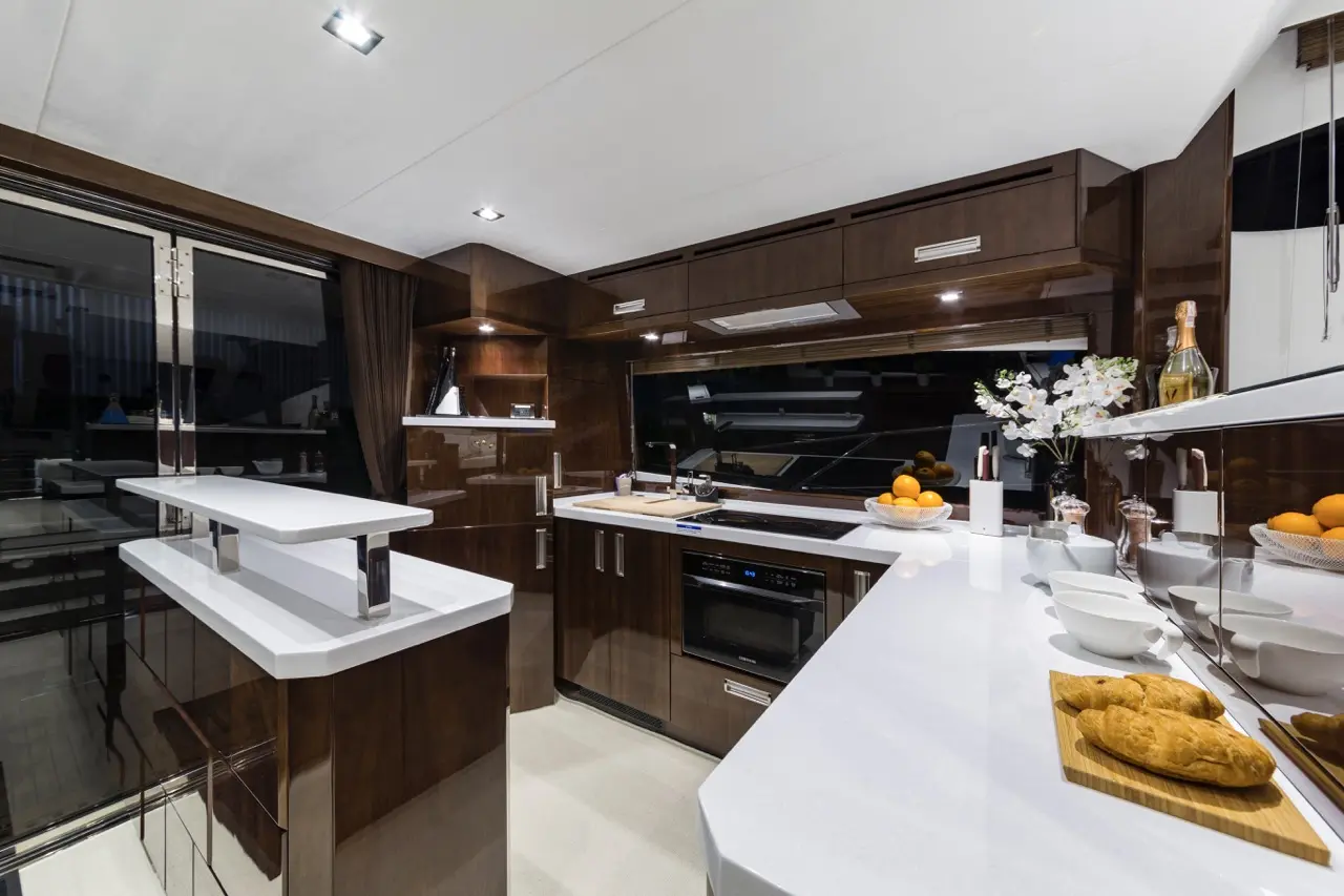 Modern yacht kitchen featuring glossy wood cabinets, white countertops, baked goods, and fresh fruit, illuminated by recessed lighting.