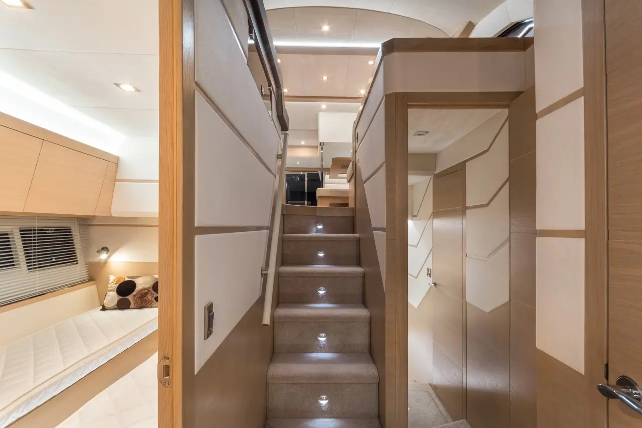 Interior view of a yacht with a staircase, soft lighting, wood paneling, and a peek into a cabin with twin beds and pillows.