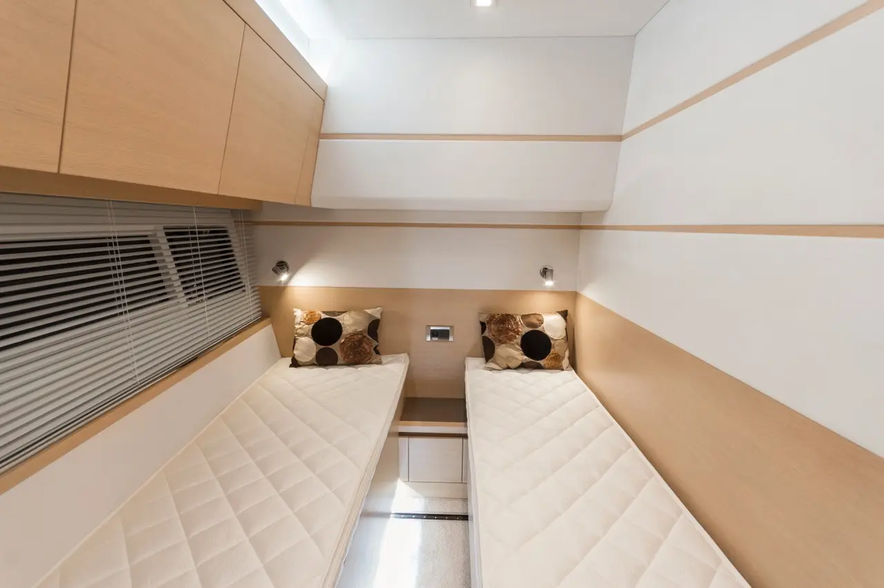 Two single beds with cream mattresses and patterned pillows in a modern room featuring wooden cabinetry and window blinds.