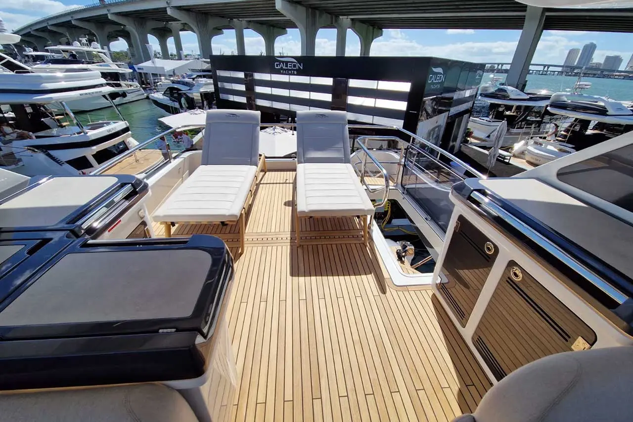 Luxurious yacht deck with two loungers, wooden flooring, and Galeon Yachts booth in the background, showcasing a marina setting.