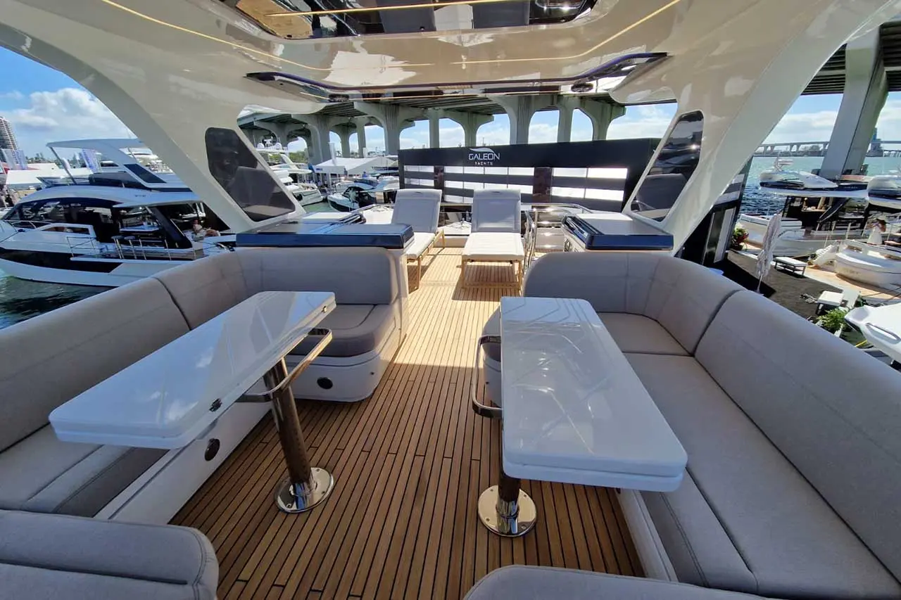 Luxurious yacht deck featuring gray seating, white tables, and views of nearby boats under a blue sky. Galeon Yachts branding visible.