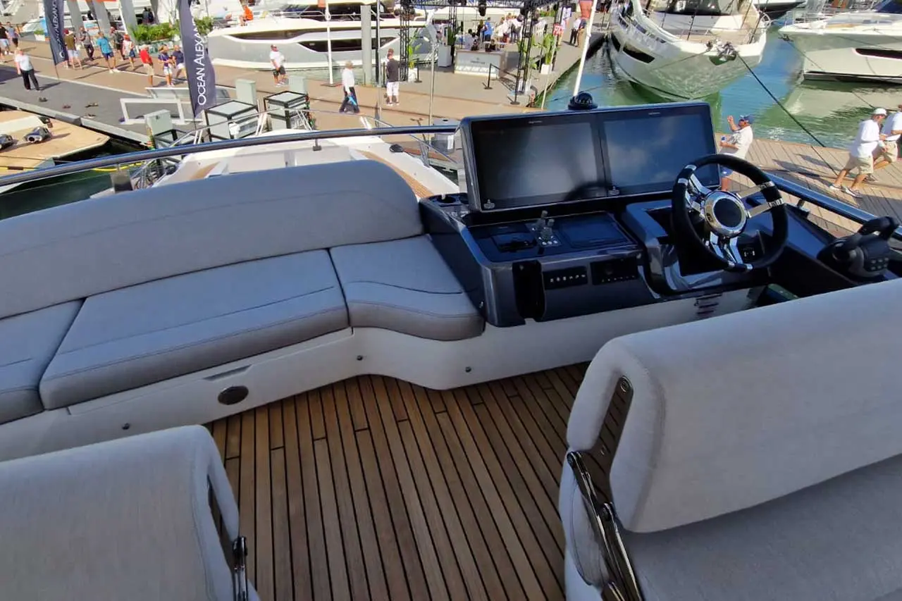 Luxury yacht cockpit features plush seating, dual screens, and steering wheel, with bustling marina in the background.