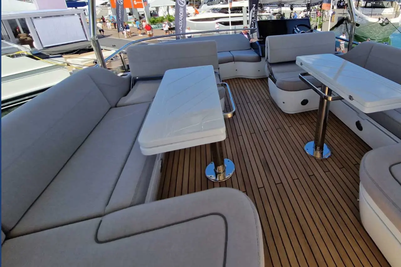 A spacious boat deck featuring a grey seating area, two tables, and a teak floor, with yachts and visitors visible in the background.