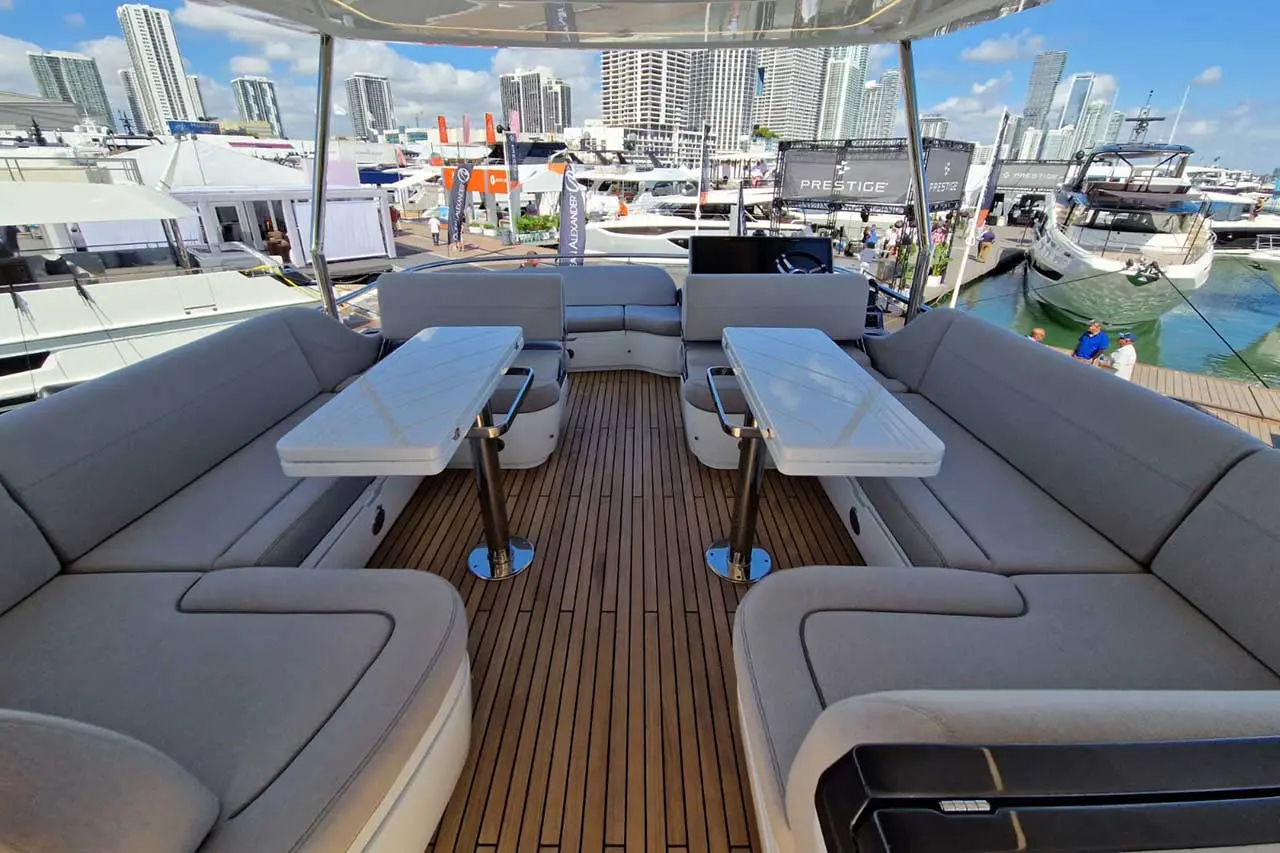 Luxury yacht interior featuring comfortable gray seating, white tables, and a view of a marina with skyscrapers in the background.