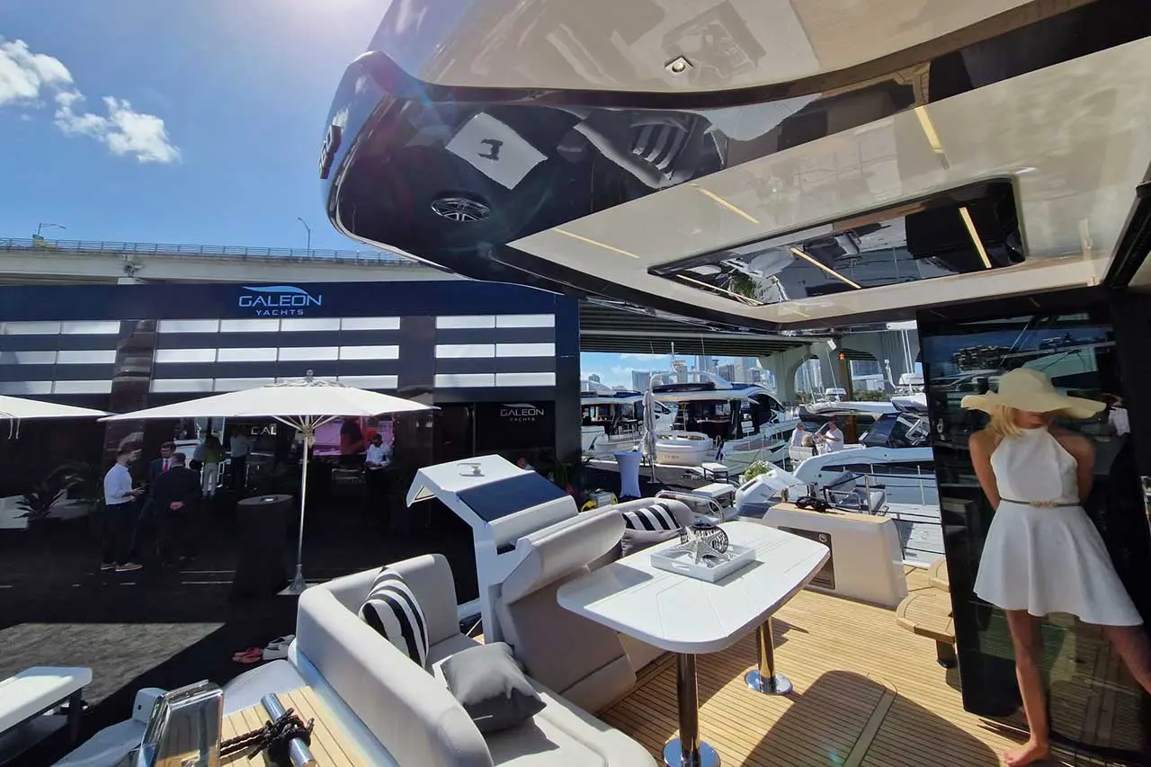 Sunlit yacht deck with sleek seating, a table, and a woman in a white dress and hat, overlooking galeon yachts display.