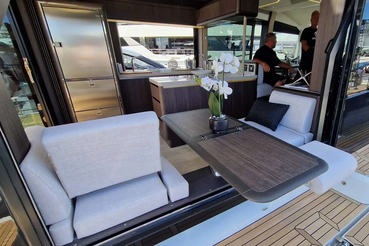 Modern yacht interior featuring a dining table, plush seating, a kitchenette, and decorative orchids. Two men are seated nearby.