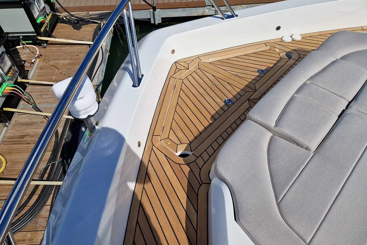 View of a boat deck featuring teak wood planks, a plush gray cushion, and a chrome railing with dock wiring visible.