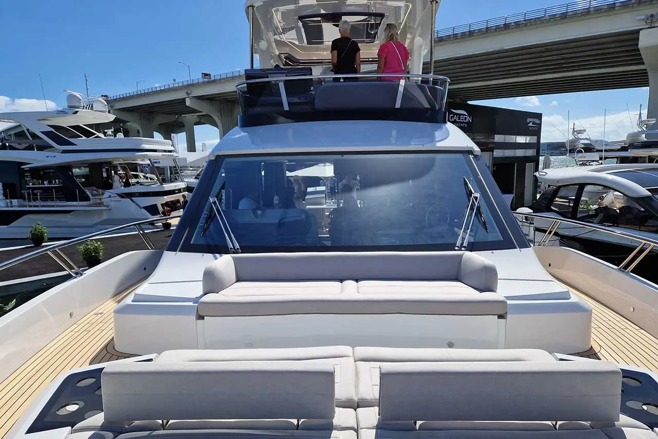 Two people on a yacht's upper deck under blue skies, with luxury boats and a concrete bridge in the background.