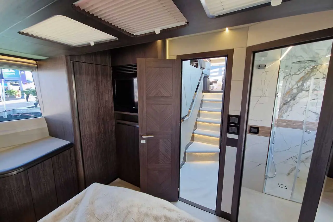 Modern yacht interior showing a bedroom with a TV, open door to a bathroom, and illuminated staircase leading up.