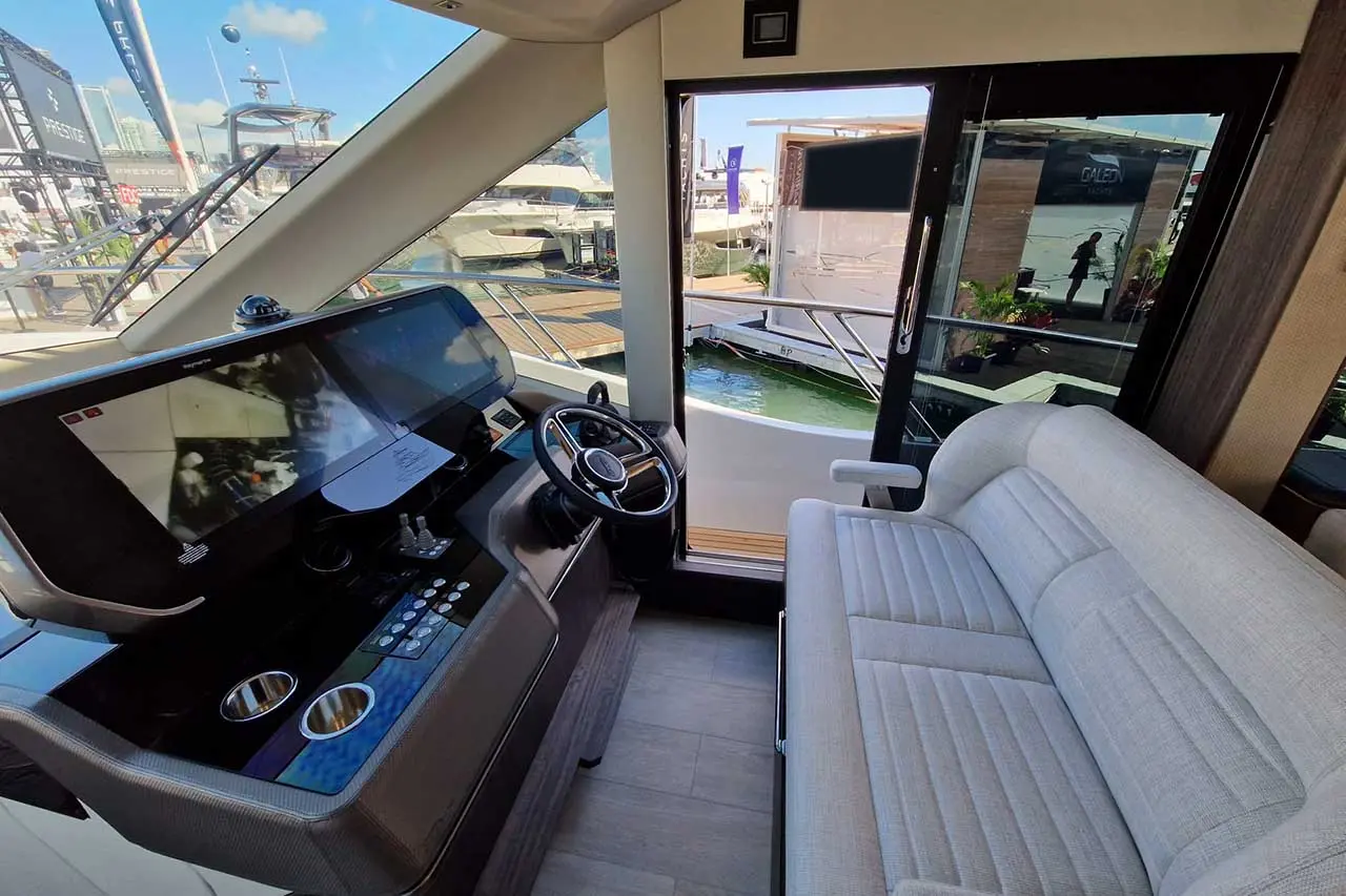 Modern yacht interior featuring dual screens, steering wheel, and a gray sofa, with a view of the marina outside.