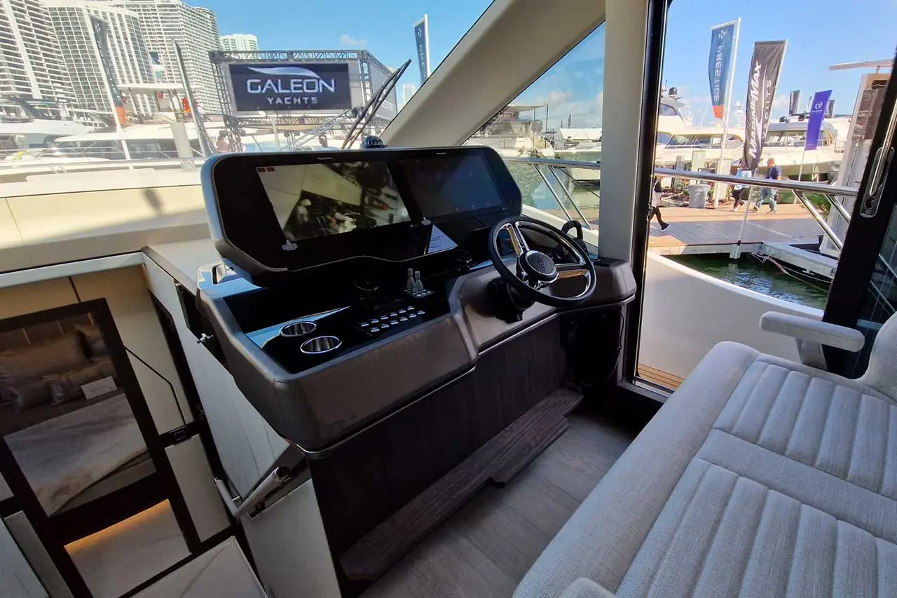Interior of a yacht's control station with dual screens, steering wheel, and view of the Galeon Yachts banner outside.