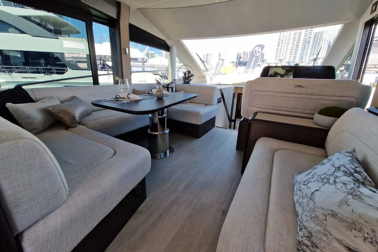 Modern yacht interior featuring a lounge area with grey seating, a dining table, and large windows overlooking a marina.