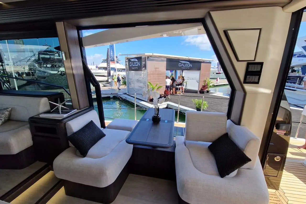 Interior view of a yacht showing modern seating, a table, and a window view of the Galeon Yachts booth at a marina.