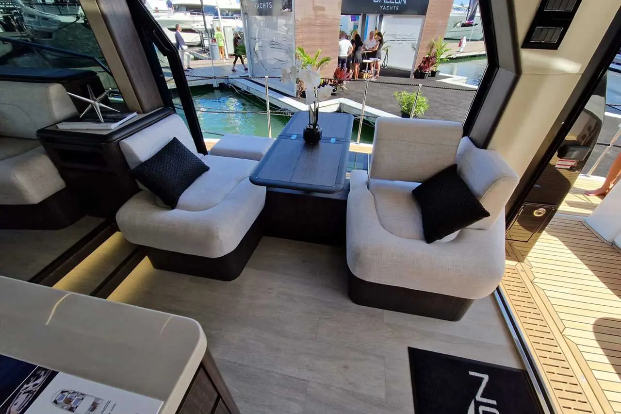 Luxurious yacht interior with white seating, black cushions, a dark wooden table, and a view of the marina outside.