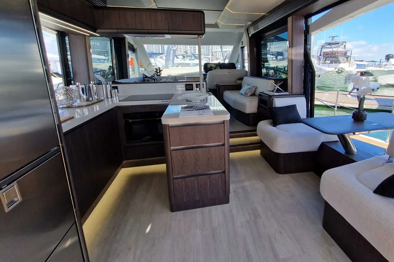 Modern yacht interior featuring a sleek kitchen, lounge area, and large windows with views of the marina and boats.