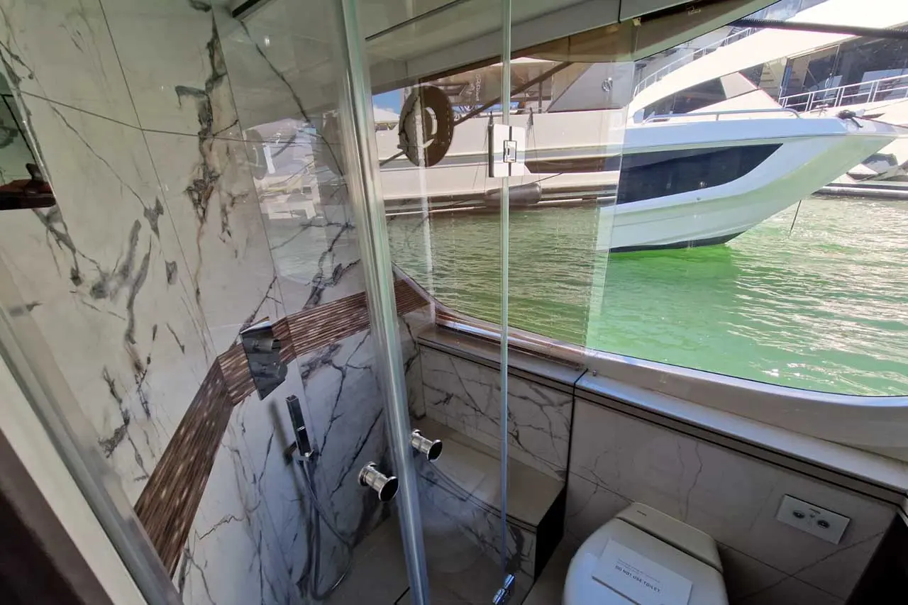 Luxurious boat bathroom featuring marble walls, a shower with glass doors, and a toilet overlooking green water outside.