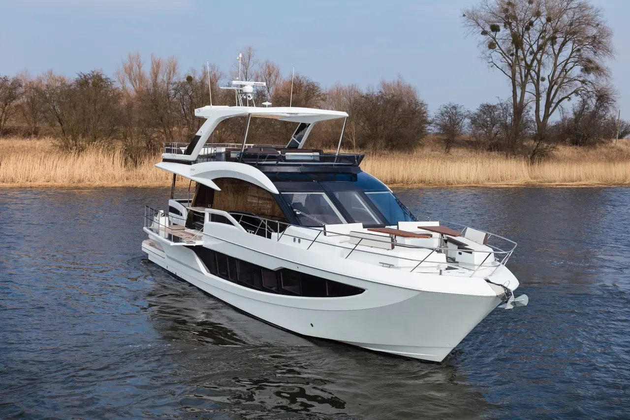 A luxurious white motor yacht navigates a serene waterway with reed banks in the background and a clear blue sky above.