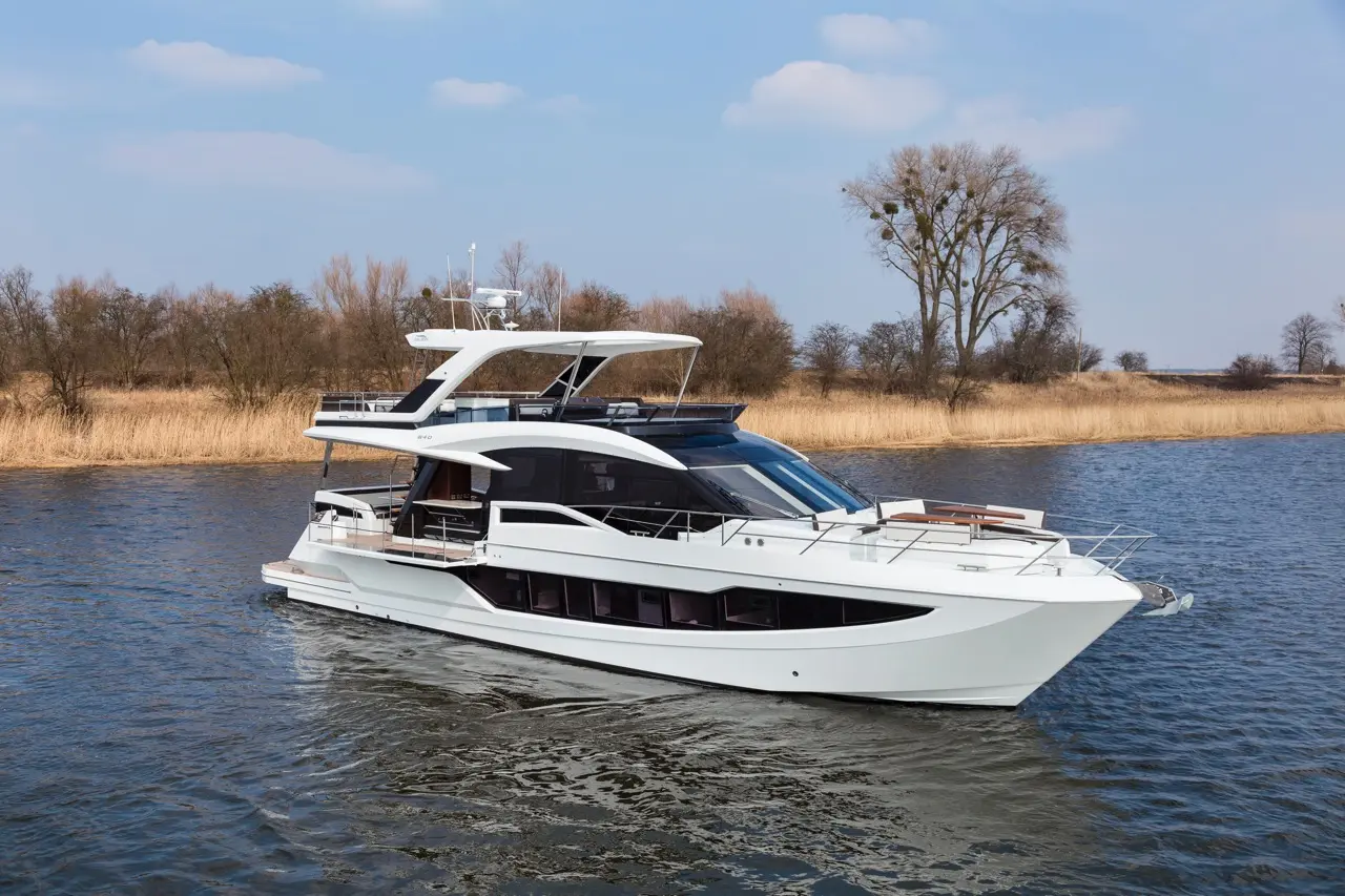 Luxury motor yacht cruising on calm water with trees and reeds in the background under a partly cloudy sky.