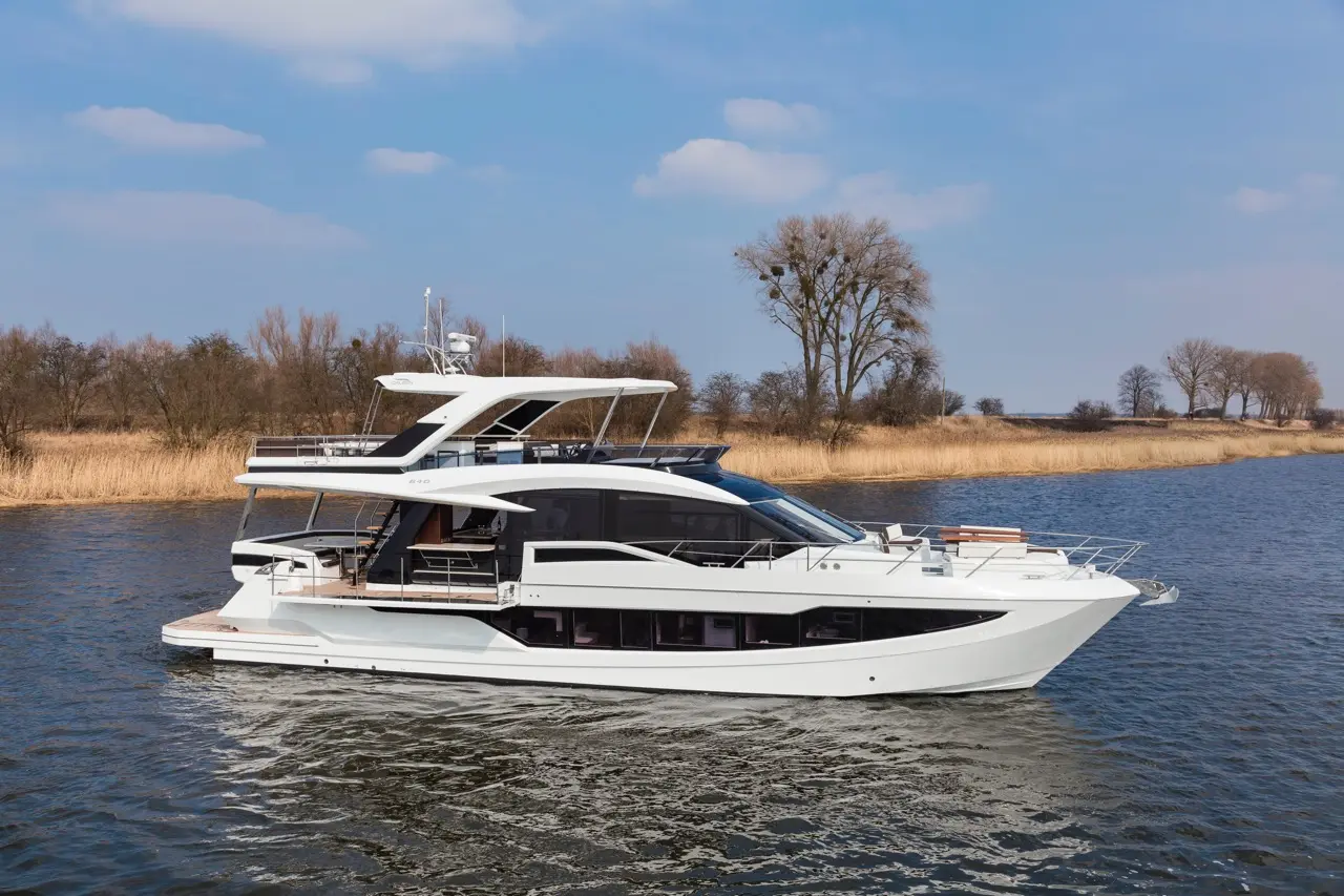 A sleek luxury yacht gliding through calm waters, surrounded by trees and golden reeds under a blue sky.