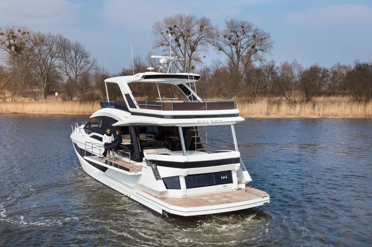 A sleek Galleon yacht navigates calm waters, with two people relaxing on the deck against a backdrop of trees.