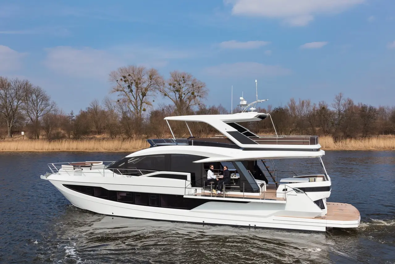 Luxury motor yacht cruising on a river, with two people onboard, surrounded by trees and blue sky.