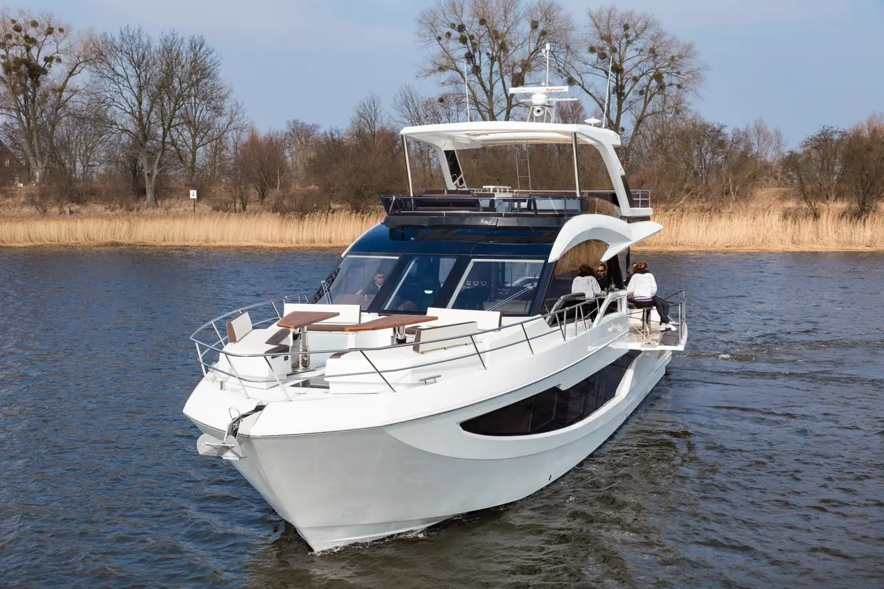 A sleek white motor yacht navigates a river, with passengers seated on deck, surrounded by trees and reeds.