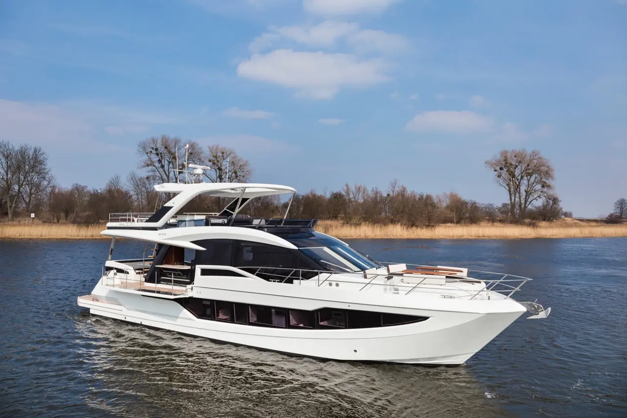 A sleek white yacht glides through calm waters, surrounded by trees and reeds under a bright blue sky.