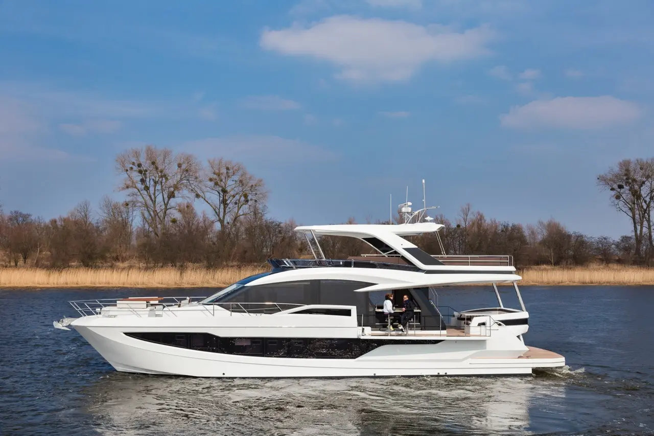 A sleek white yacht cruising on calm water, with trees and dry grass in the background under a bright blue sky.