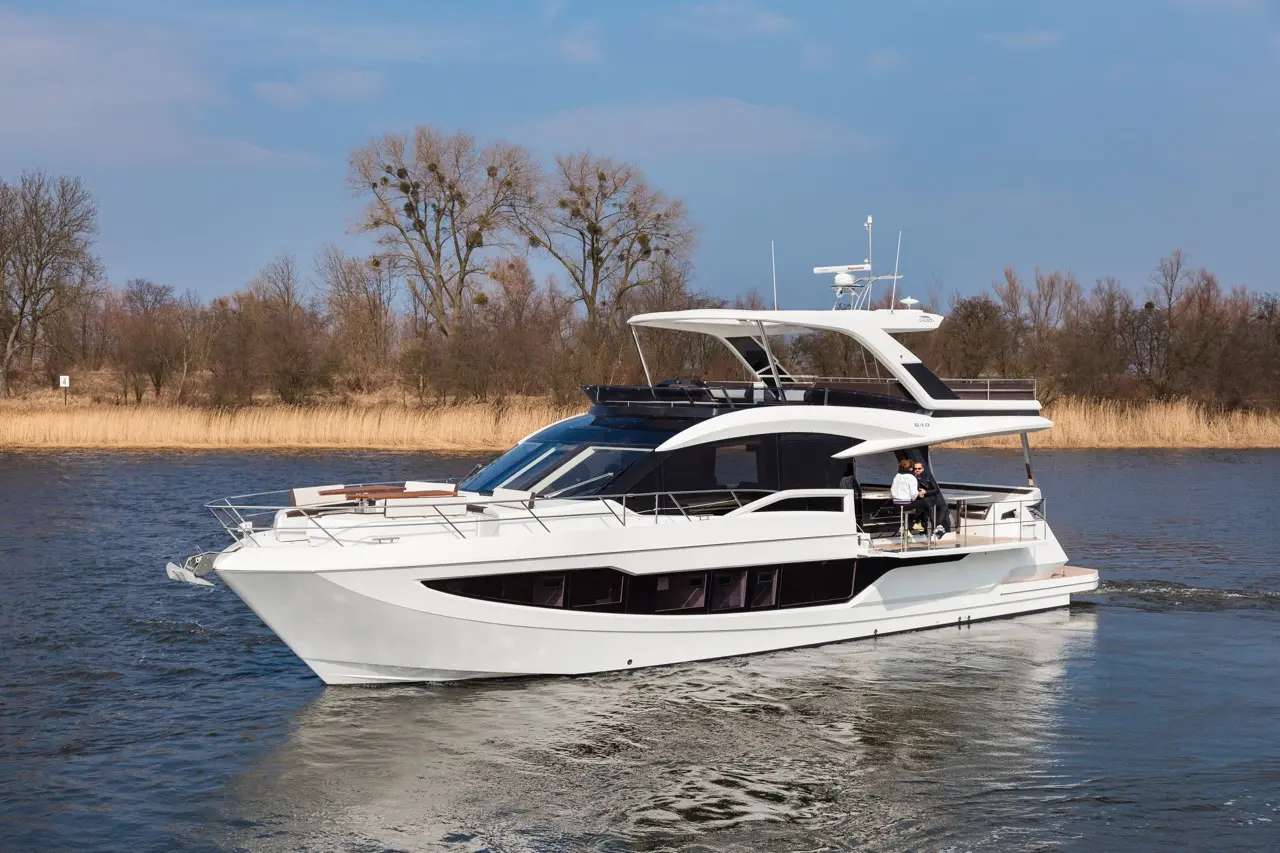 A sleek white yacht cruising on calm waters, with two people enjoying the view on the deck under clear blue skies.