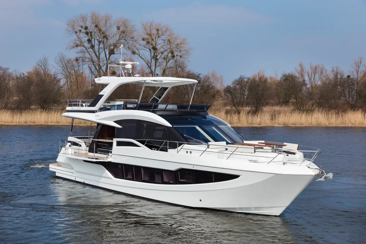A sleek white motor yacht cruising on calm water, surrounded by trees and dry grasses against a clear blue sky.